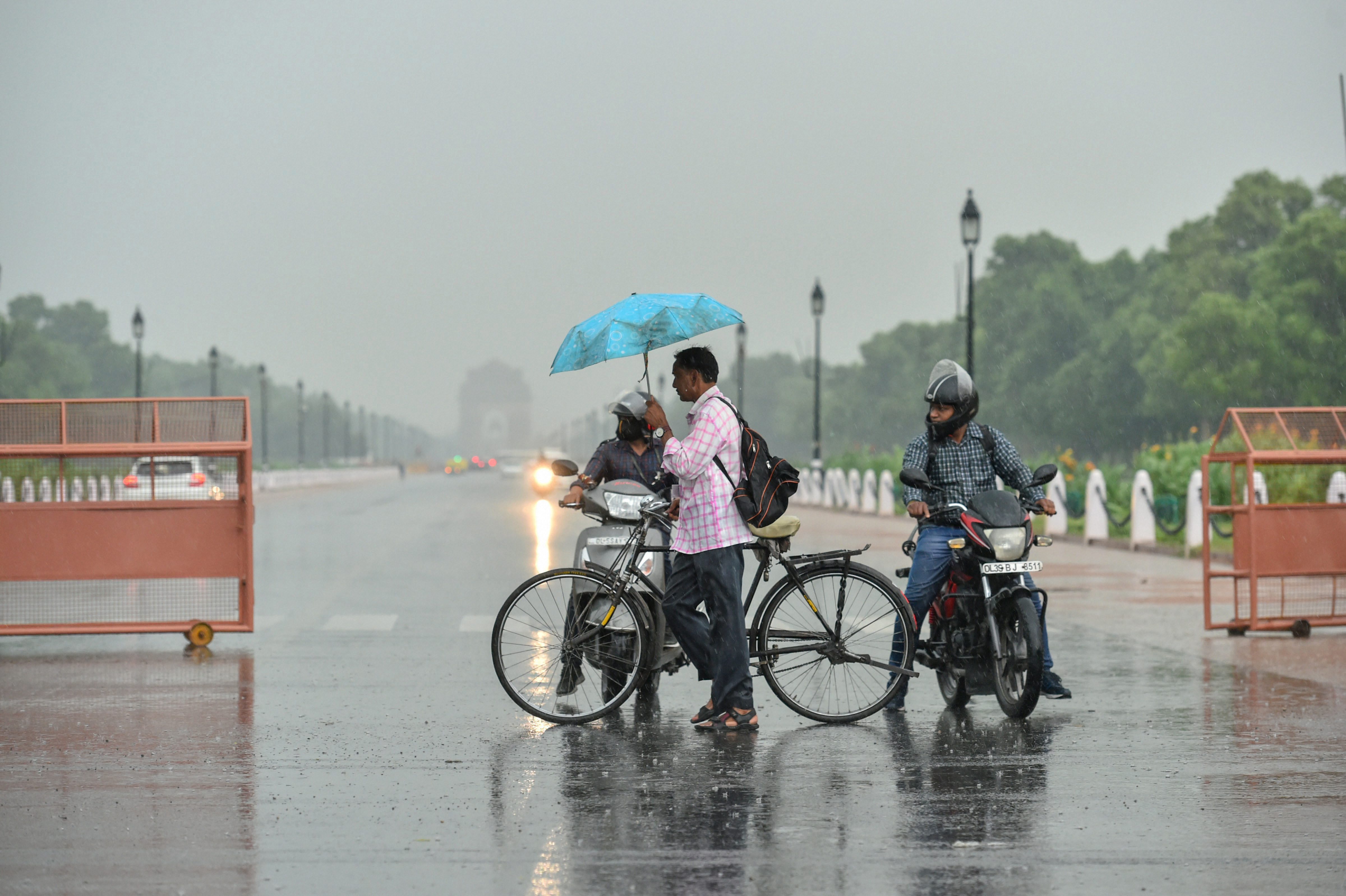 A cyclist holding an umbrella crosses a road during rainfall, in New Delhi - PTI